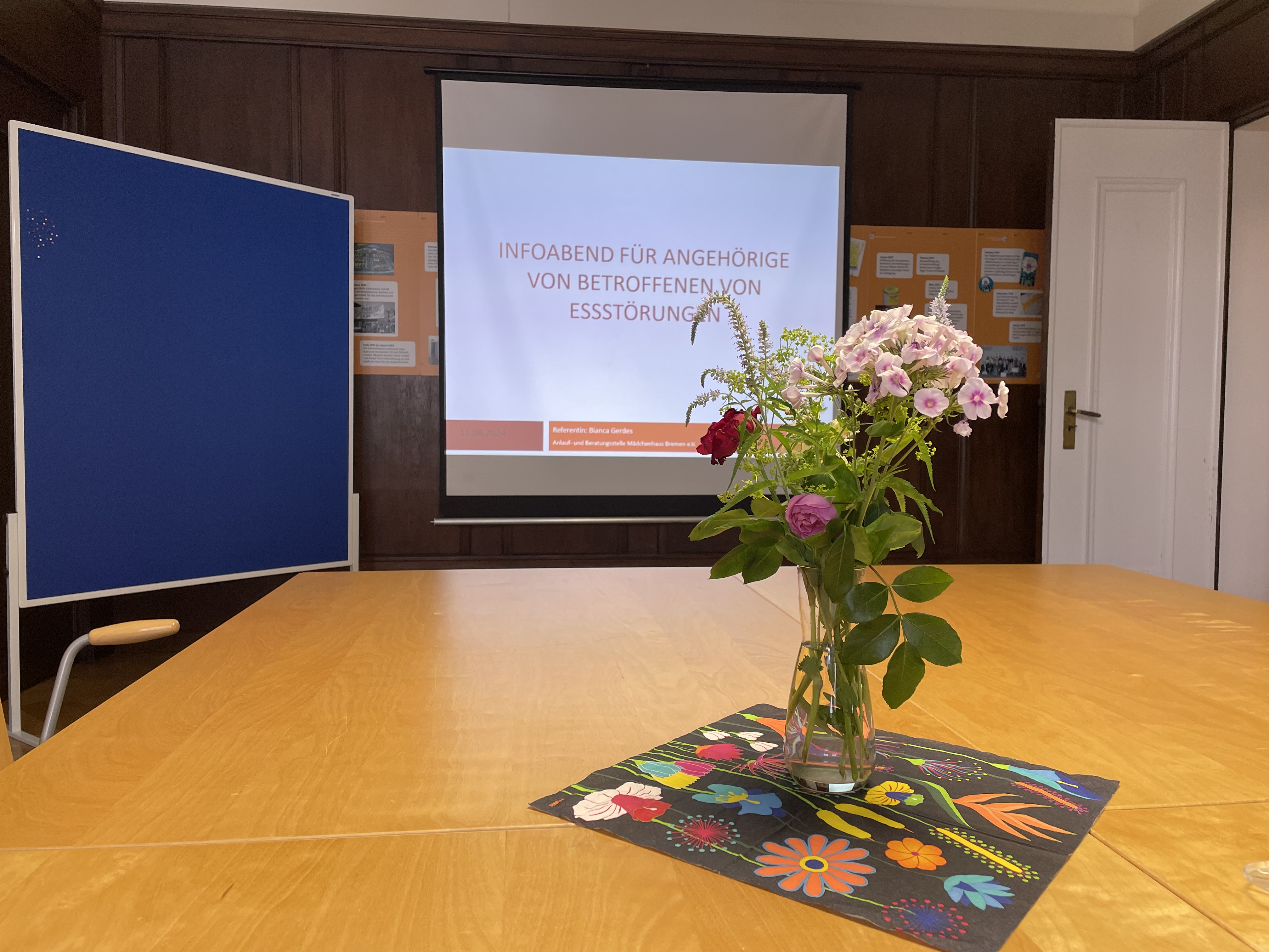 Auf dem Bild ist ein Tisch mit Blumen abgebildet. Im Hintergrund hängt eine Leinwand mit dem Titel: "Infoabend für Angehörige von Betroffenen von Essstörungen".